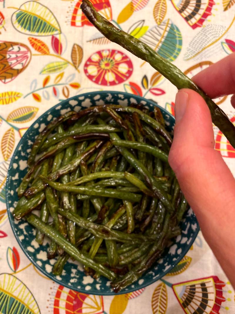 woman holding a roasted green bean with several other roasted green beans in a teal and white bowl underneath