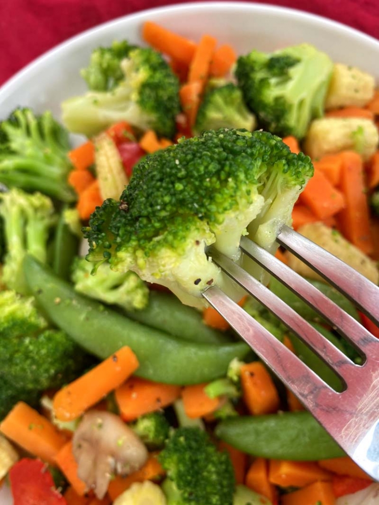 A fork with a piece of broccoli on it, over a bowl of mixed vegetables 