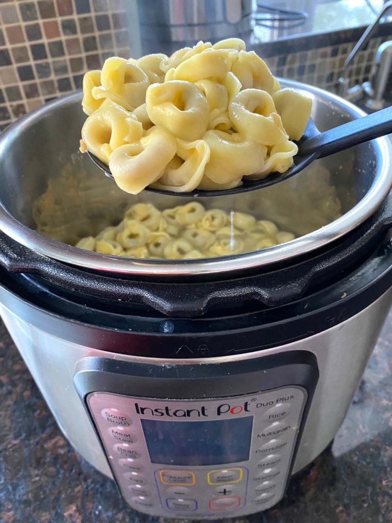 A spoon of cooked tortellini being held over an instant pot full of cooked tortellini.