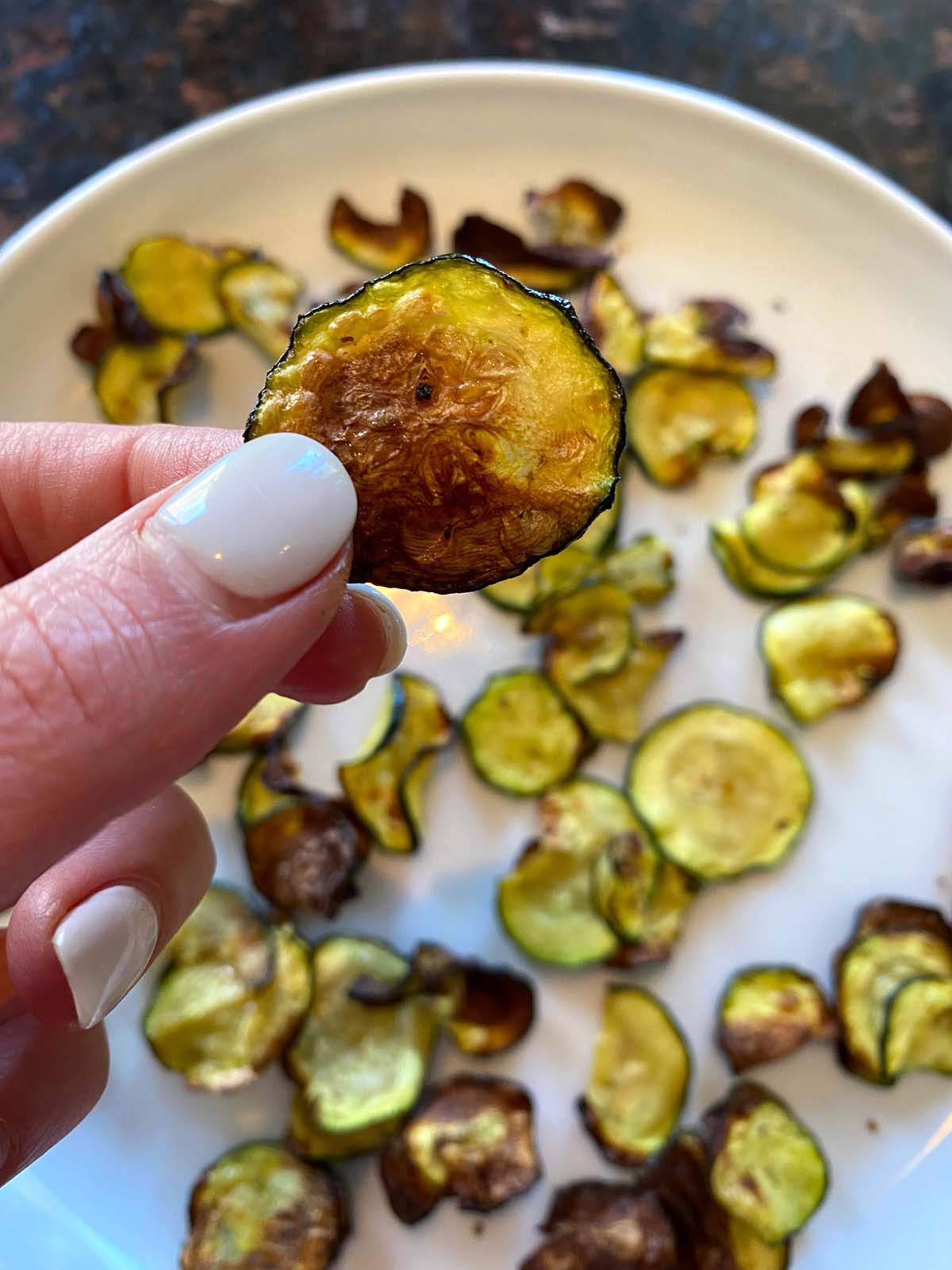 Hand holding a zucchini chip up close with plate of chips underneath.