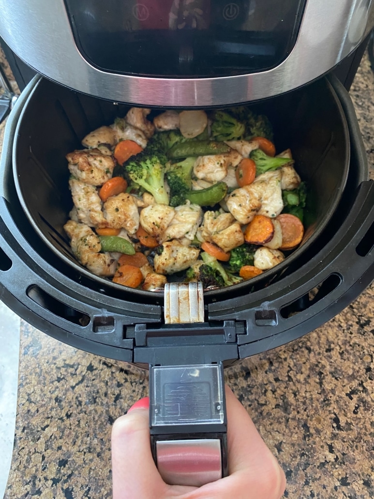 hand opening air fryer to show chicken and vegetables cooking inside