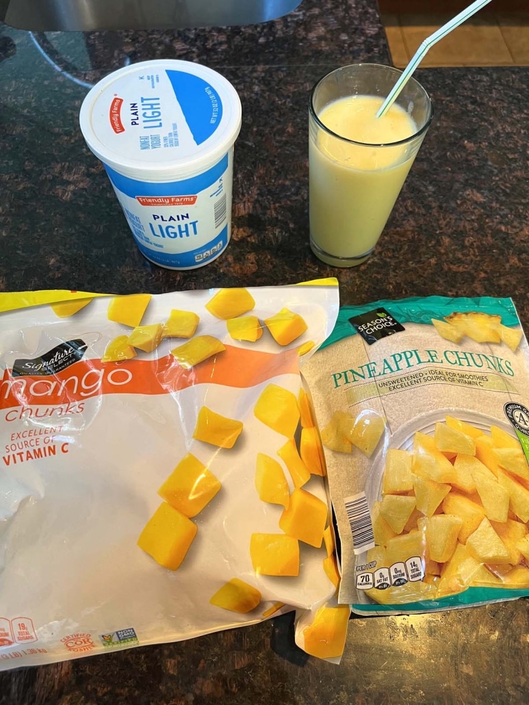 glass of Mango Pineapple Smoothie next to a container of plain yogurt and frozen fruit chunks