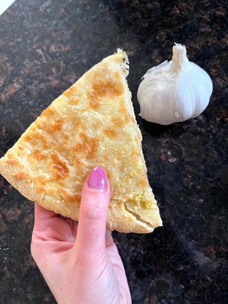 hand holding a slice of Garlic Pizza next to a garlic clove