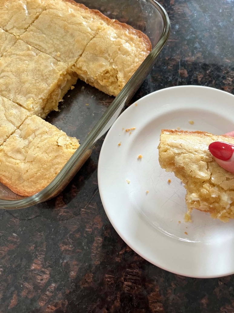 hand placing Vanilla Brownies With White Chocolate Chips on plate next to a pan of vanilla brownies