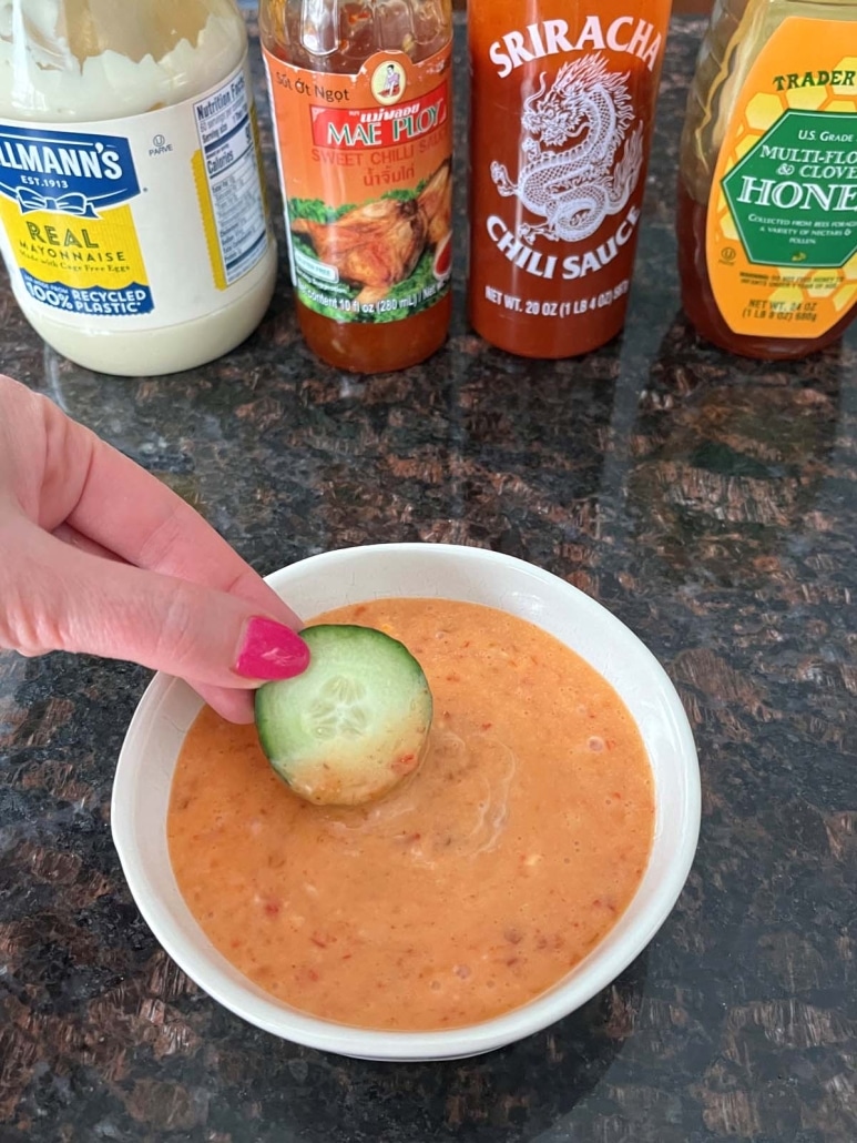 hand dipping a slice of cucumber into bang bang sauce, with mayo, sweet chili sauce, sriracha, and honey behind the bowl