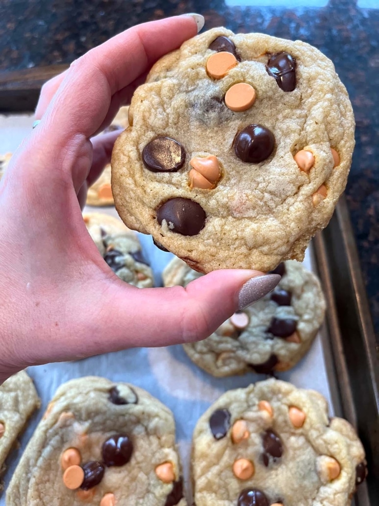 hand holding a Caramel Chocolate Chip Cookie in front of a baking sheet of cookies