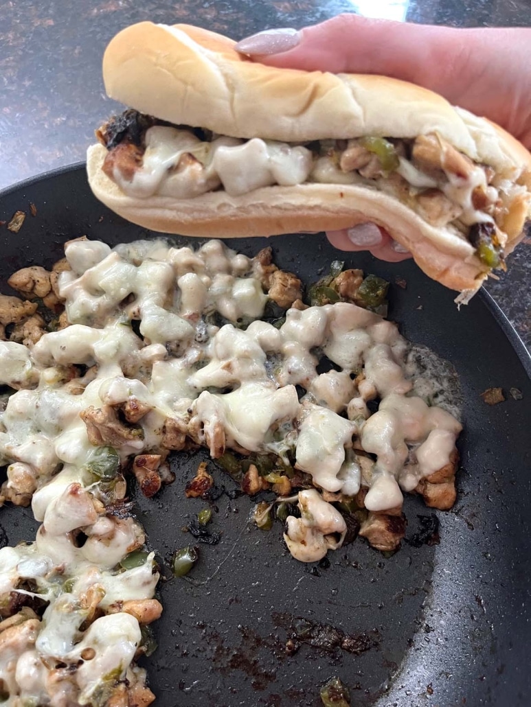 hand holding Chicken Philly Cheesesteak next to a skillet of chicken cheesesteak