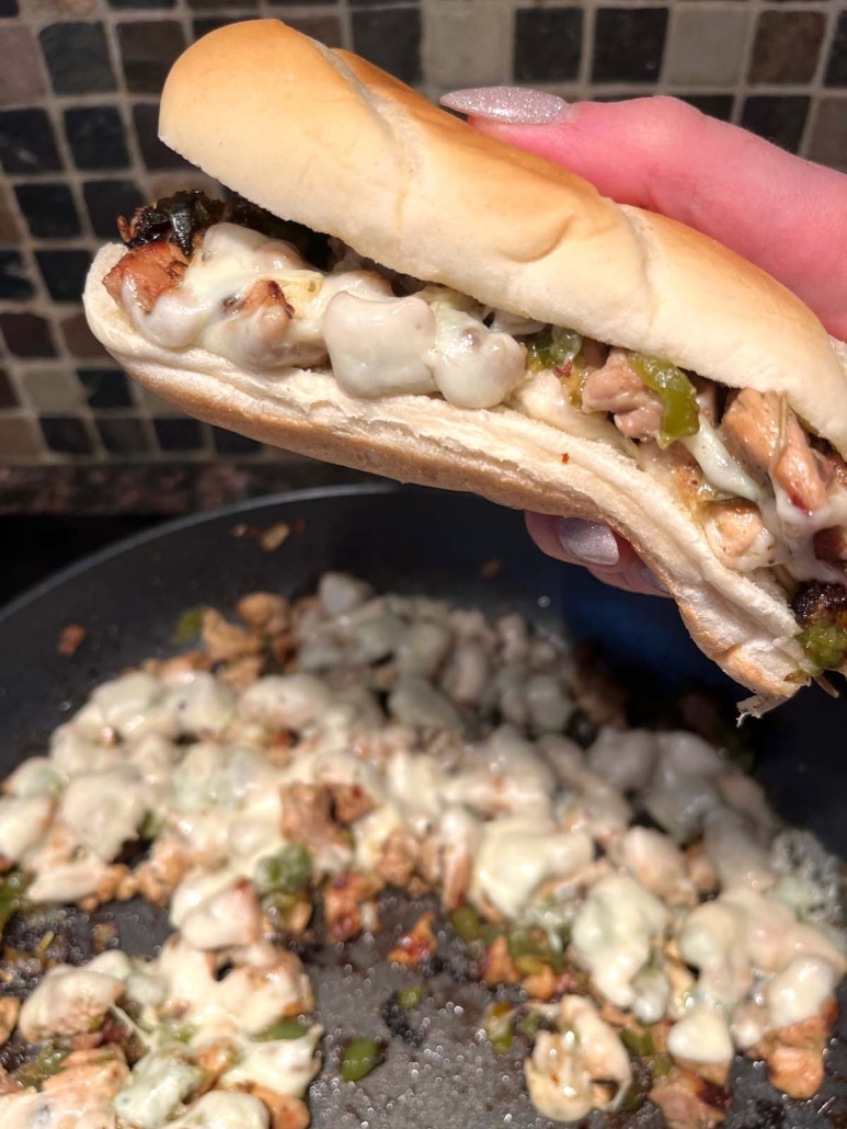 hand holding a Chicken Philly Cheesesteak above a skillet with chicken and cheese