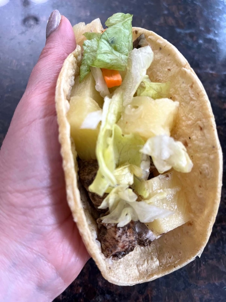 hand holding Jerk Chicken Taco, topped with sauce, lettuce, and pineapple