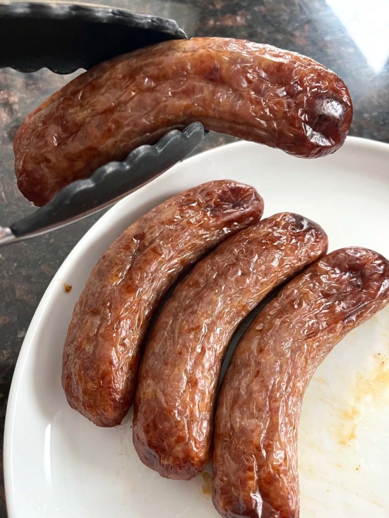 tongs holding a brat above a plate of brats