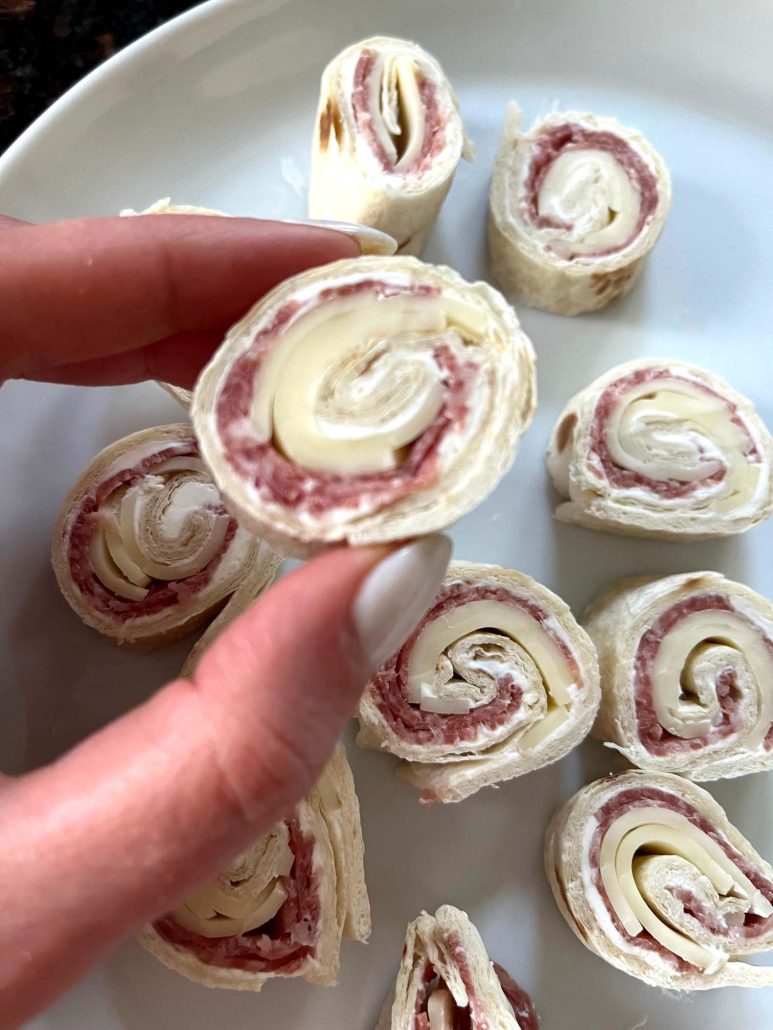 hand holding a Italian Pinwheel With Salami And Cream Cheese in front of a plate of pinwheels