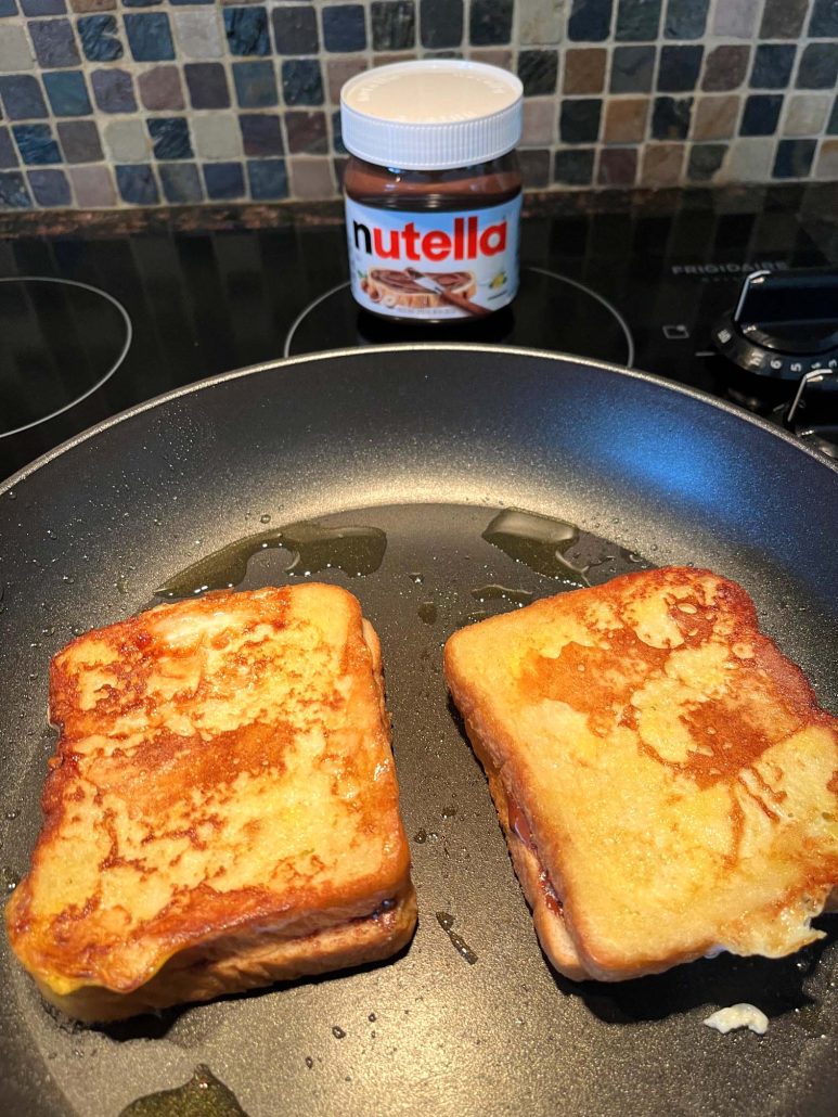 Nutella Stuffed French Toast cooking up golden brown on skillet next to a jar of Nutella