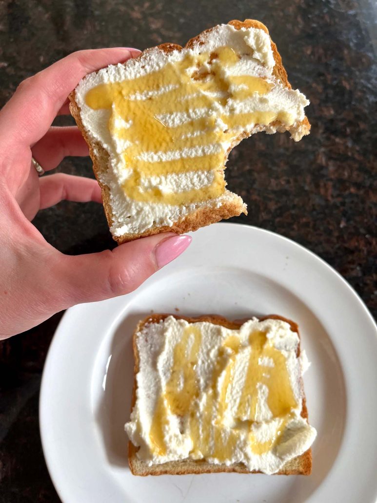 hand holding slice of Ricotta Toast with bite take out, next to another slice of toast on a plate