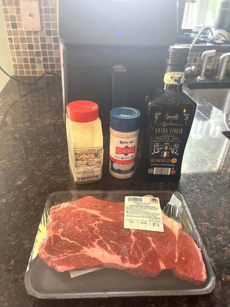 Ingredients for air fryer boneless chuck steak, including a pack of chuck steak, extra virgin olive oil, salt, and black pepper, placed in front of an air fryer.