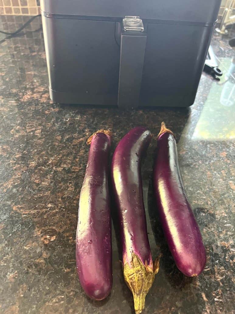 Three whole Chinese eggplants placed on a kitchen countertop in front of an air fryer, ready to be prepared for an air fryer recipe.