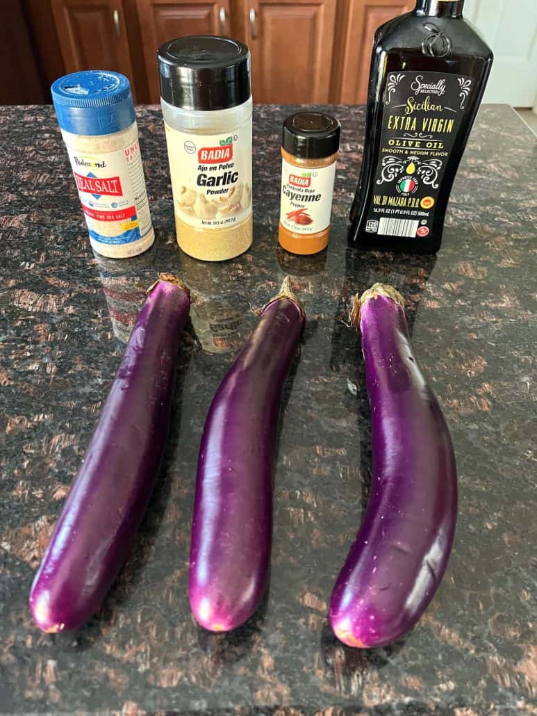 Three fresh Chinese eggplants on a countertop alongside key ingredients for seasoning: salt, garlic powder, cayenne pepper, and extra virgin olive oil, ready to be used in an air fryer recipe.