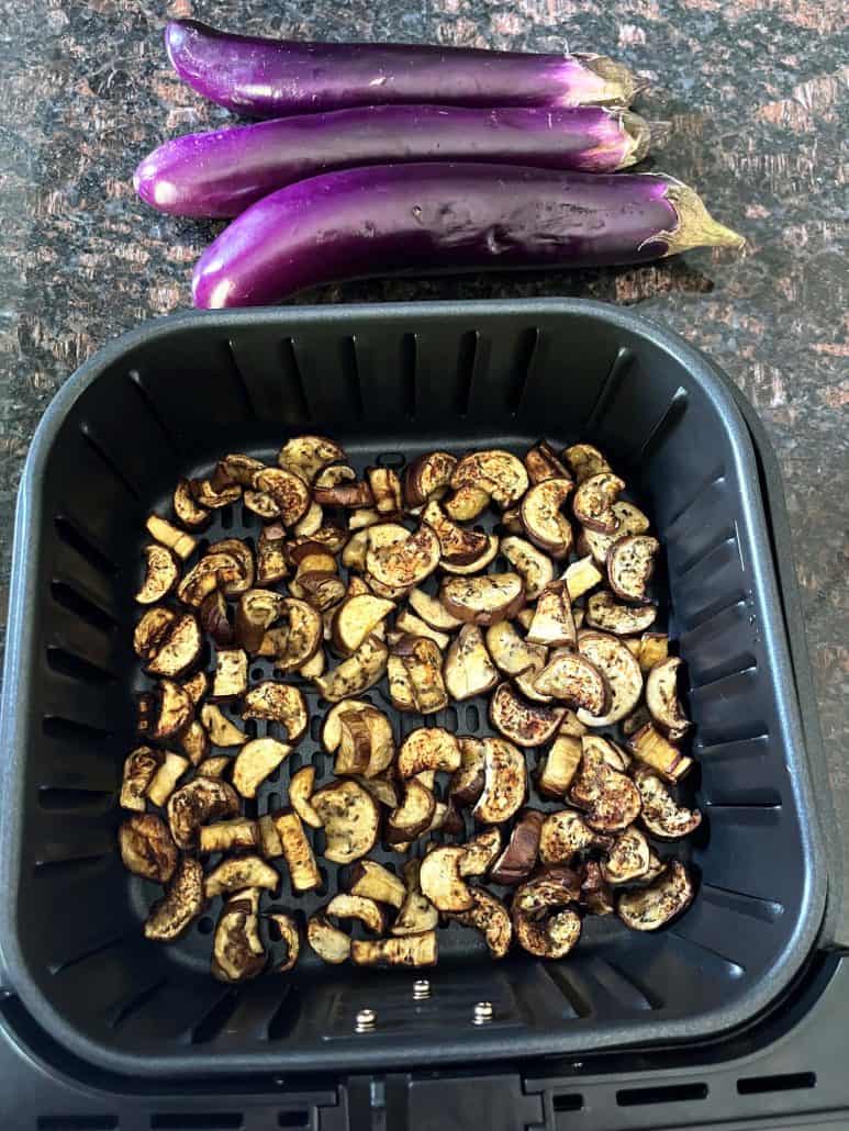 Three fresh Chinese eggplants next to an air fryer basket filled with crispy, roasted eggplant chunks, showcasing the preparation and cooking process for a healthy air fryer recipe.