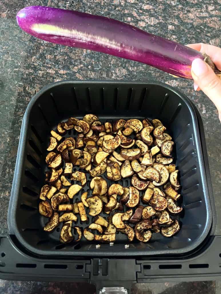 A hand holding a fresh Chinese eggplant above an air fryer basket filled with golden, crispy eggplant chunks, illustrating the ingredients and cooking process for an air fryer recipe.