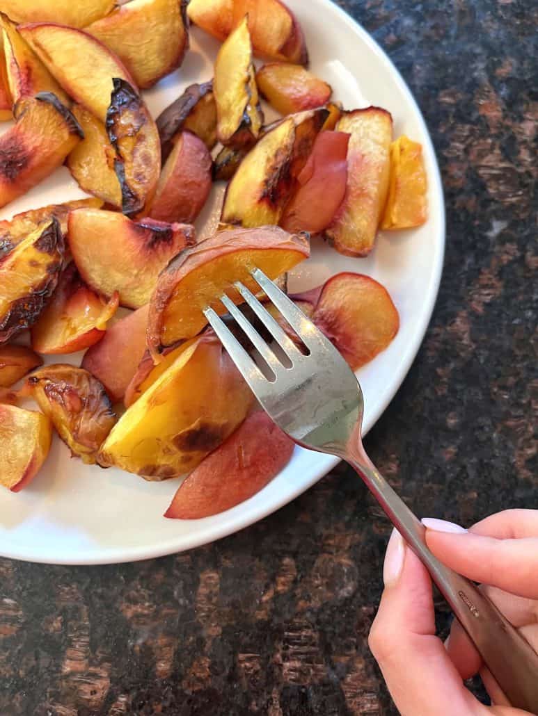 A fork holding a slice of roasted peach above a plate filled with air fryer roasted peaches.