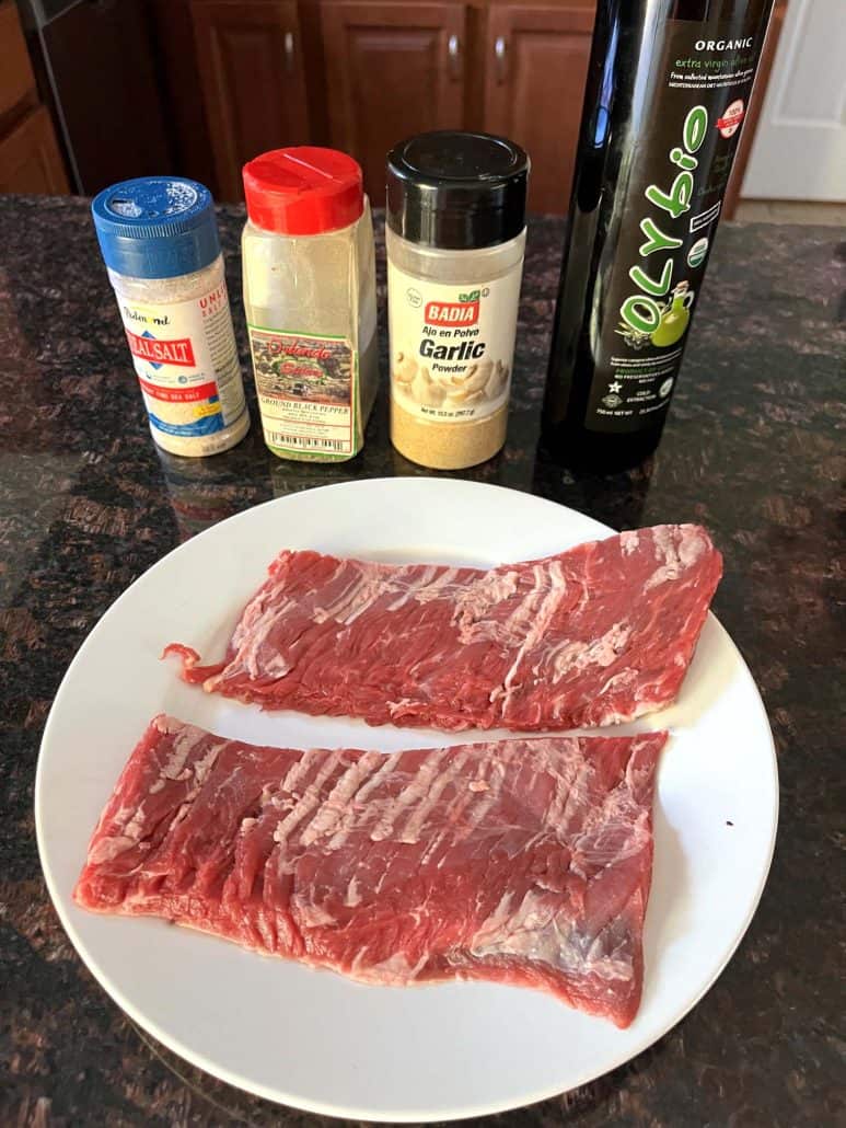 Two raw skirt steaks on a white plate with salt, pepper, garlic powder, and olive oil in the background, ready to be seasoned for the air fryer skirt steak recipe.