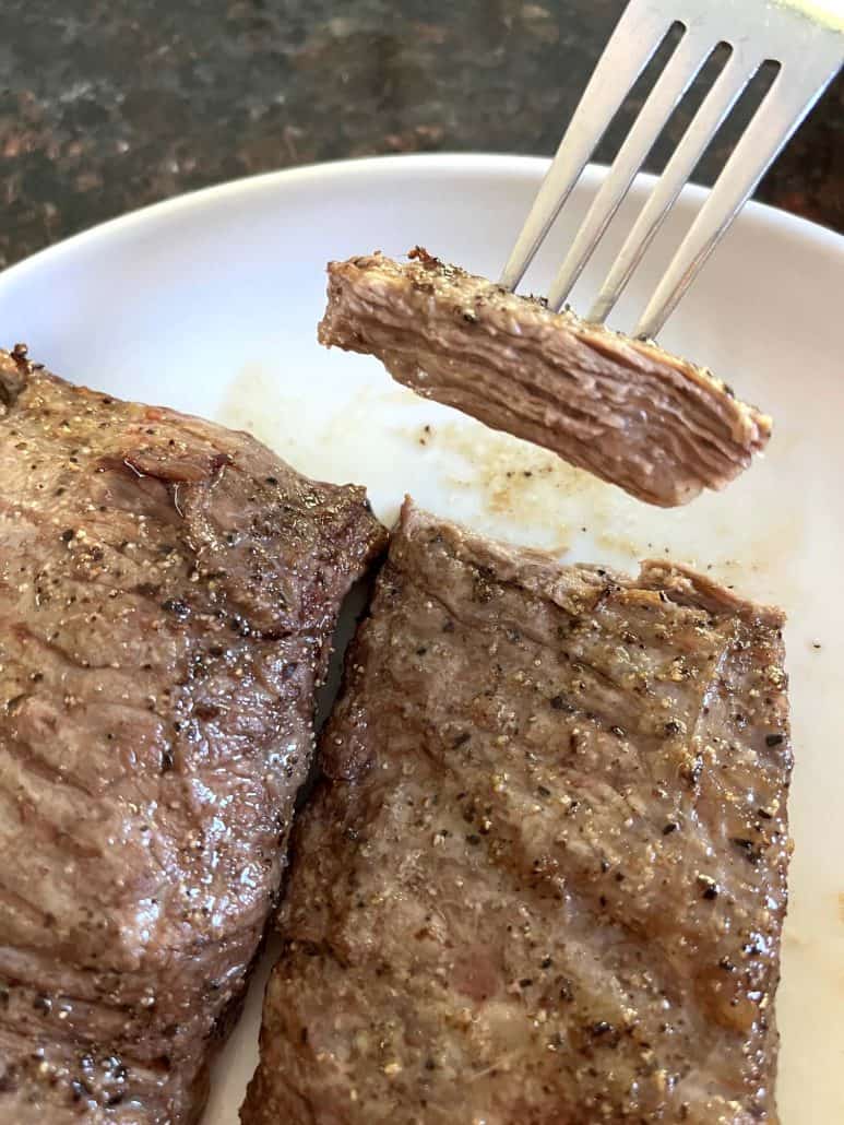 A close-up of a fork holding a slice of tender skirt steak above the rest of the steak on a white plate, showing the juicy interior after air frying.