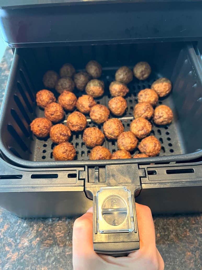 Hand holding the handle of an air fryer basket filled with crispy, golden-brown vegan meatballs fresh out of the air fryer.