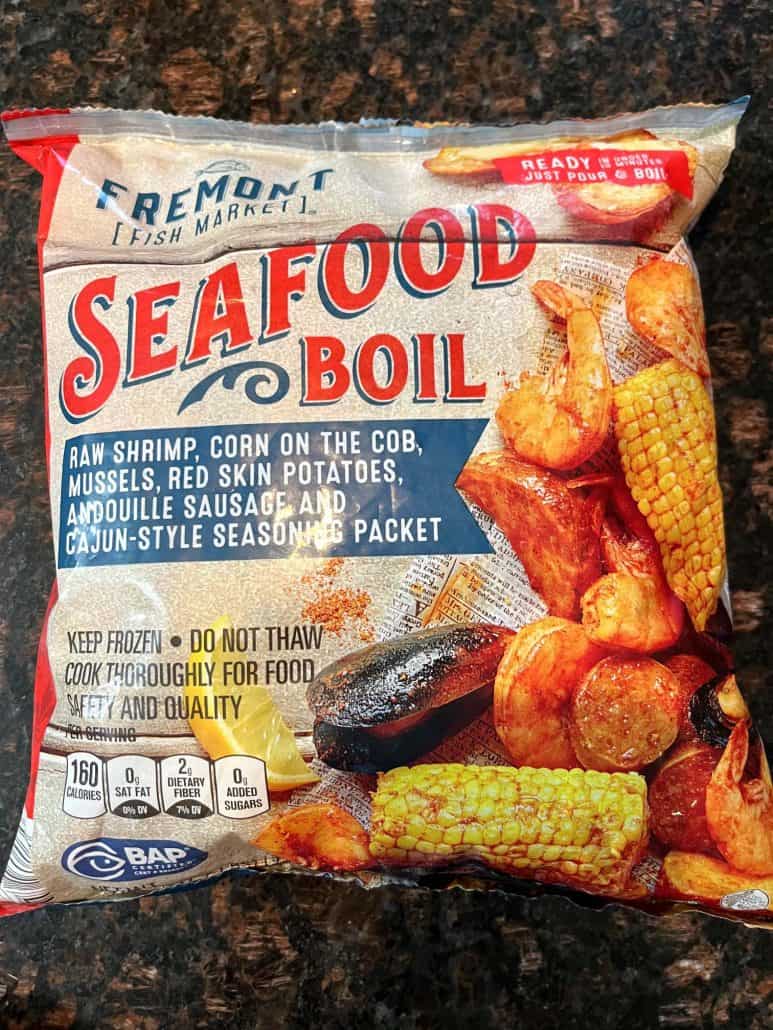 A package of Aldi's Fremont Fish Market Frozen Seafood Boil featuring raw shrimp, corn on the cob, mussels, red skin potatoes, andouille sausage, and a Cajun-style seasoning packet.