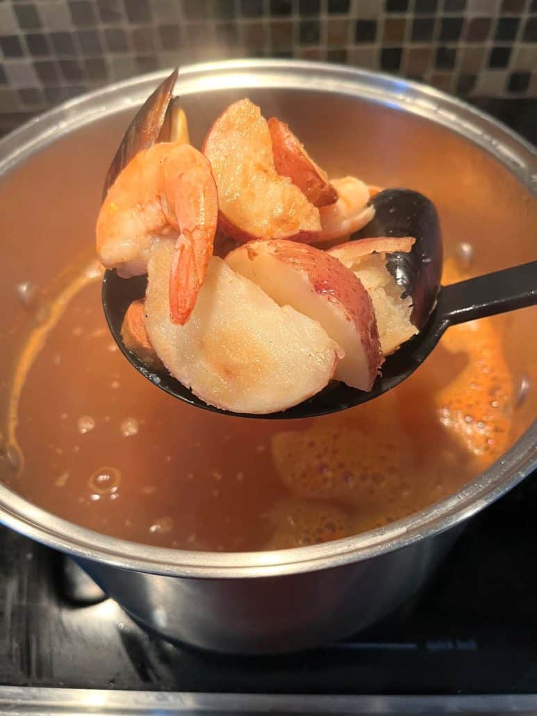 A close-up of a spoon lifting shrimp, red potatoes, and mussels from a pot of boiling Cajun-seasoned broth, as part of the cooking process for Aldi's Frozen Seafood Boil.