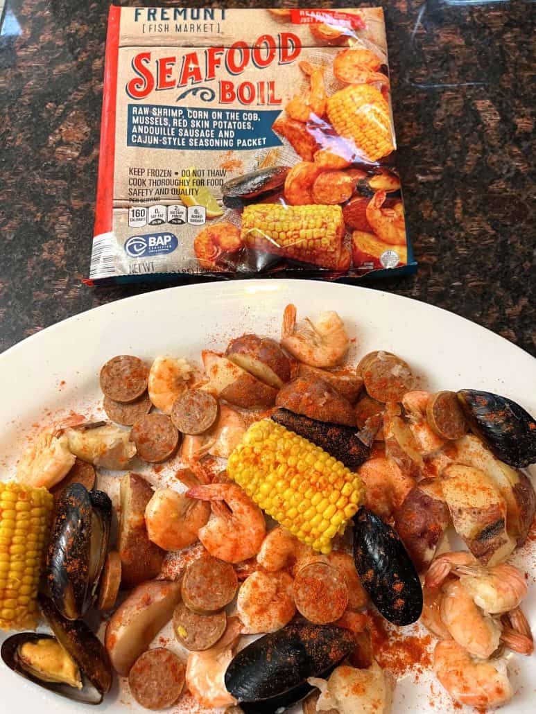 A plate of cooked shrimp, mussels, sausage, red potatoes, and corn on the cob from Aldi's Frozen Seafood Boil is displayed in front of the product's packaging.