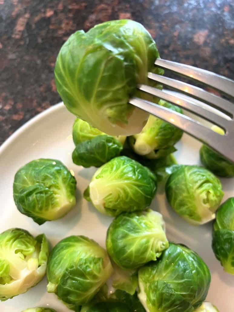 A fork holding a boiled Brussels sprout above a plate filled with more vibrant green sprouts.