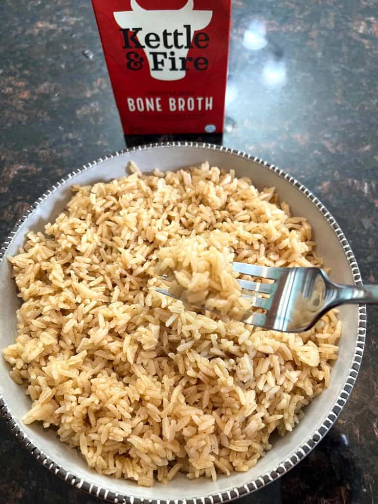 A fork lifting a portion of fluffy bone broth rice from a bowl, with a package of Kettle & Fire Bone Broth in the background.