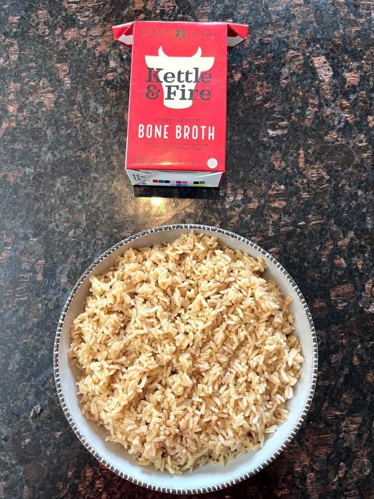 A bowl of cooked bone broth rice placed on a countertop with a package of Kettle & Fire Chipotle Beef Bone Broth displayed above it,