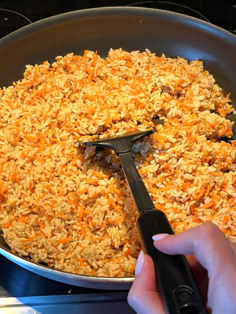 Cooking Carrot Rice in a pan, showing a hand of Melanie from melaniecooks.com stirring the mixture of cooked rice and shredded carrots, evenly sautéing.