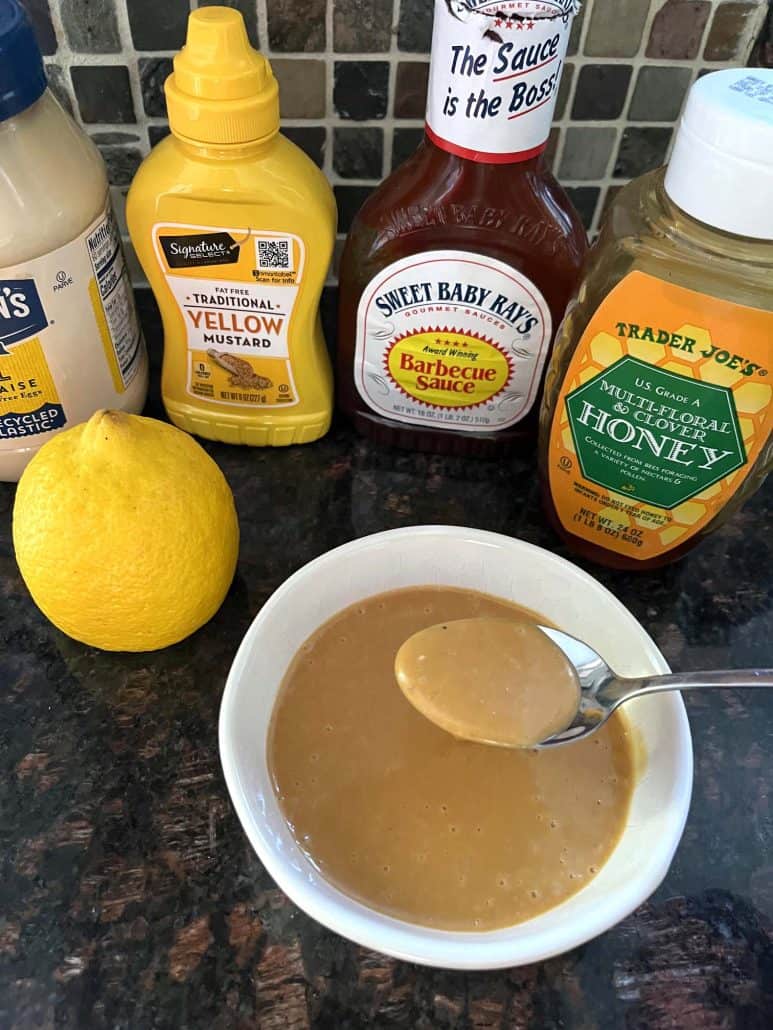 A bowl of homemade Chick-Fil-A sauce with a spoon, surrounded by the key ingredients: mayonnaise, mustard, barbecue sauce, honey, and a lemon.