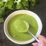 A spoon scooping green Cilantro Chimichurri from a small white bowl, with fresh cilantro leaves in the background, showcasing the sauce's smooth texture and fresh ingredients.