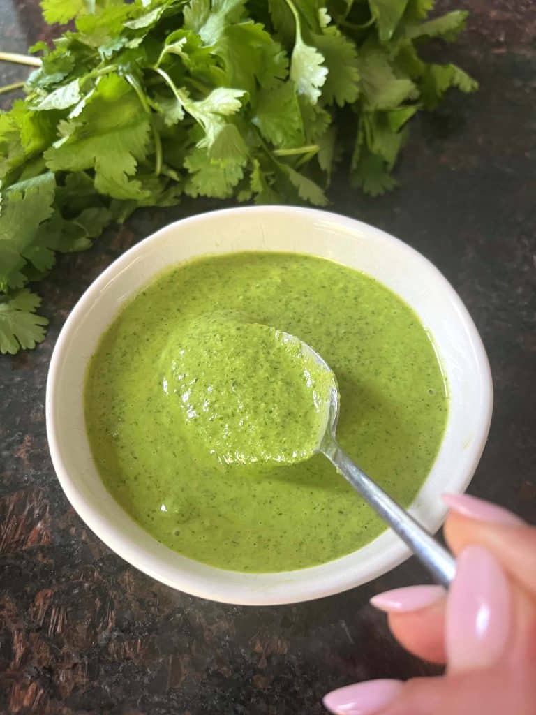 A spoon scooping green Cilantro Chimichurri from a small white bowl, with fresh cilantro leaves in the background, showcasing the sauce's smooth texture and fresh ingredients.