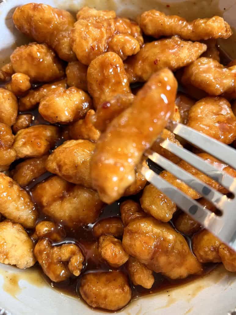 A fork lifts a piece of crispy General Tso’s chicken coated in a sticky, flavorful sauce from a bowl filled with air-fried chicken pieces.