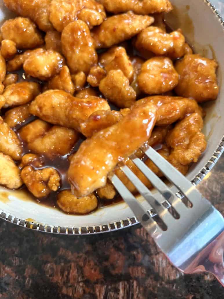 A fork holds a piece of saucy General Tso’s chicken over a bowl filled with crispy, air-fried chicken coated in a rich, flavorful sauce.