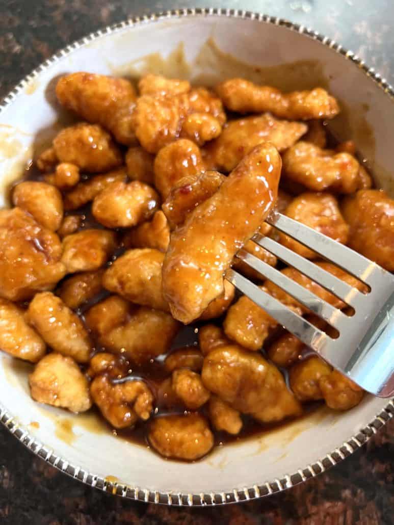 A bowl of air-fried General Tso’s chicken coated in a rich sauce, with a fork holding up a piece, showing the crispy texture and sticky glaze of the dish.