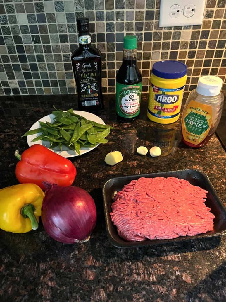 Ingredients for Ground Beef Stir Fry laid out on a kitchen counter, including a tray of raw ground beef, red and yellow bell peppers, a red onion, fresh snow peas, garlic cloves, ginger, soy sauce, cornstarch, honey, and extra virgin olive oil. 