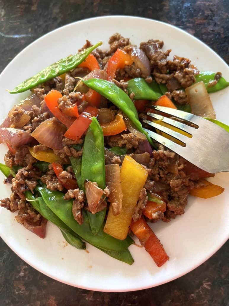 A serving of Ground Beef Stir Fry on a white plate, featuring a colorful mix of browned ground beef, red and yellow bell peppers, red onions, and crisp snow peas, with a fork resting on the side.