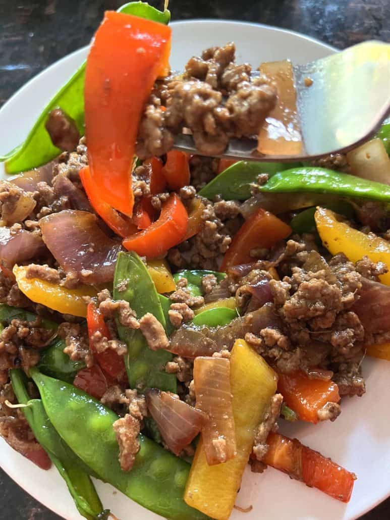 A close-up of a plate filled with a vibrant Ground Beef Stir Fry, showcasing tender ground beef, crisp red and yellow bell peppers, red onions, and green snow peas, with a fork lifting a bite.