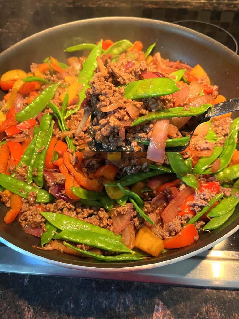 A skillet filled with Ground Beef Stir Fry, featuring a vibrant mix of ground beef, snow peas, red and yellow bell peppers, and red onions, with a spatula lifting a generous serving. 