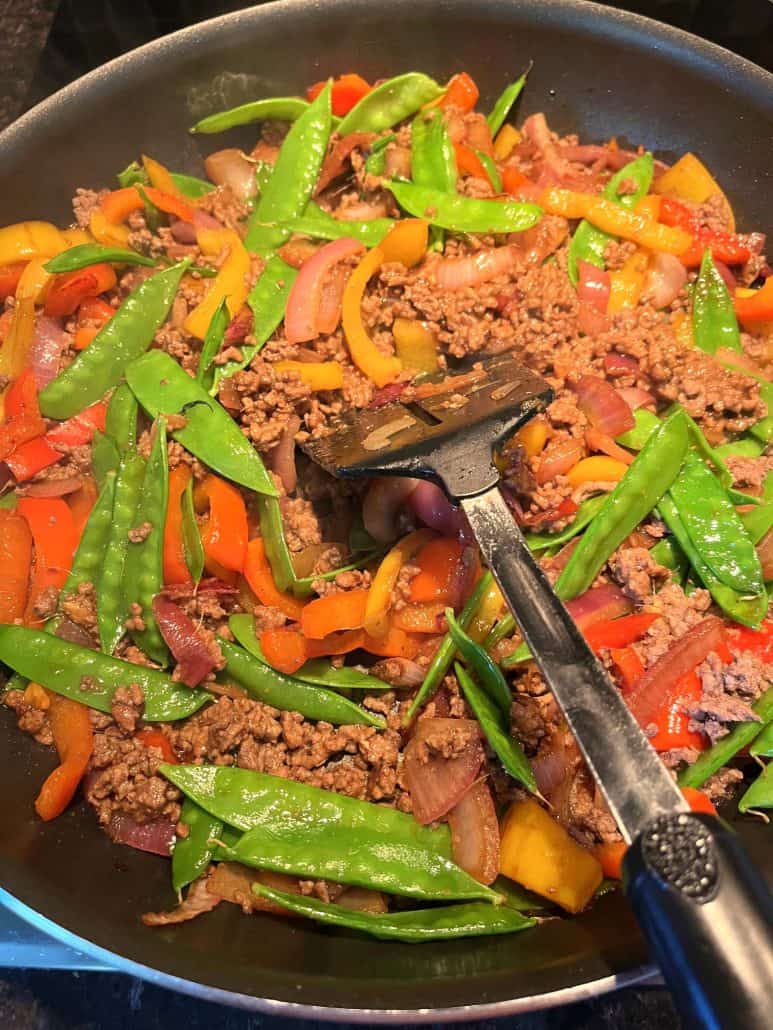  A colorful skillet filled with a freshly made Ground Beef Stir Fry, featuring browned ground beef, bright orange and red bell peppers, sliced red onions, and vibrant green snow peas, being stirred with a spatula. 