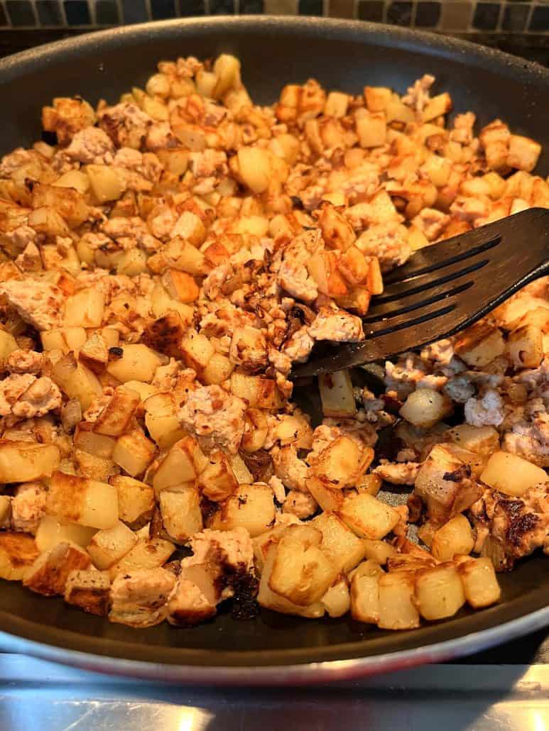 A skillet filled with browned diced potatoes and ground turkey being stirred with a spatula.