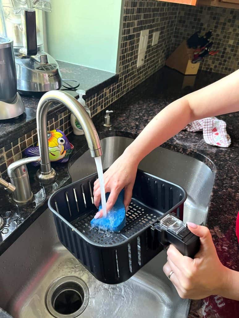 Demonstrating the hand washing a Cosori air fryer basket with a non-abrasive sponge under running water.