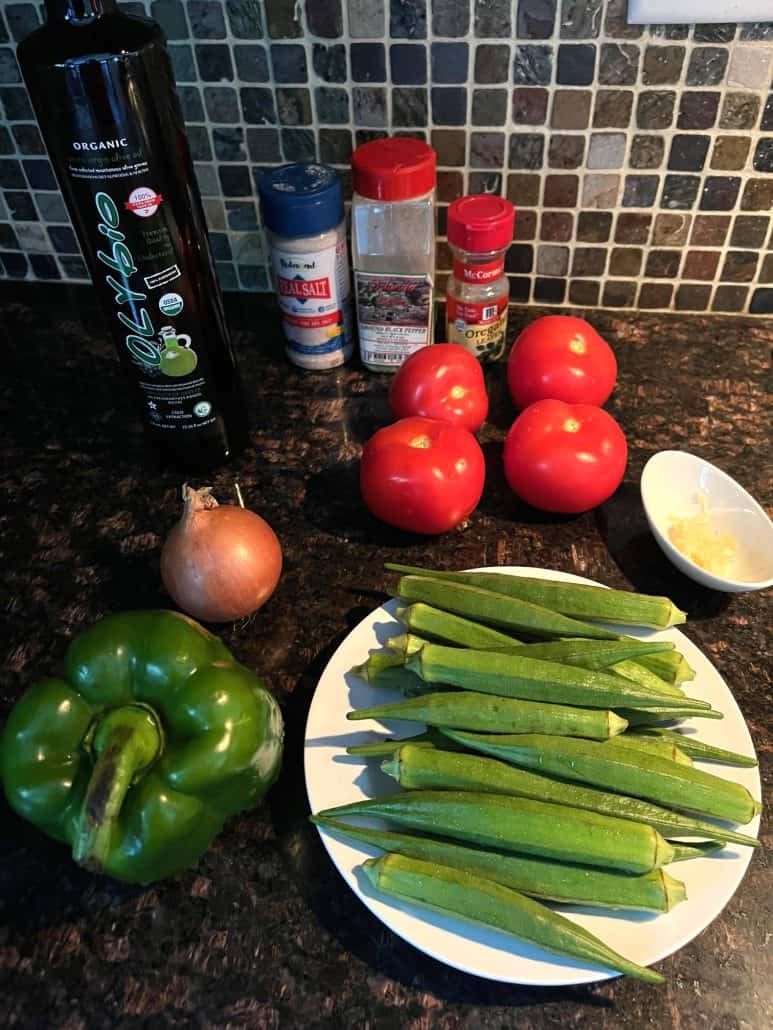 Ingredients for Okra and Tomatoes recipe laid out on a countertop, including fresh okra, tomatoes, bell pepper, onion, garlic, olive oil, and seasonings like salt, pepper, and oregano. 