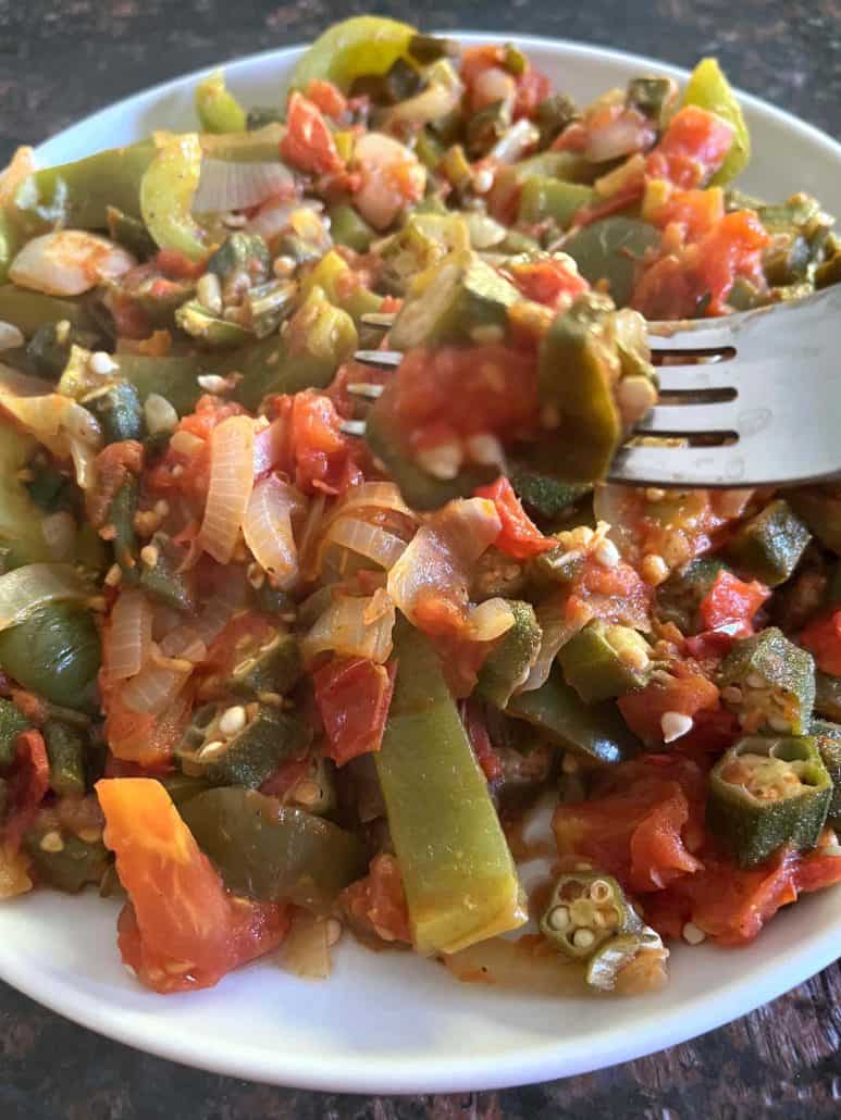 A fork lifting a bite of sautéed okra, tomatoes, bell peppers, and onions from a plate.