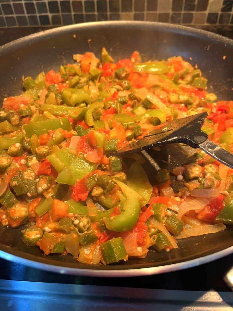 A pan filled with sautéed okra, tomatoes, bell peppers, and onions being stirred with a spatula.