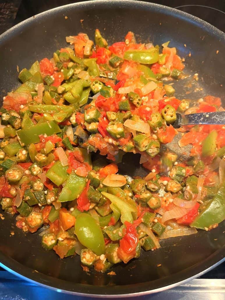 A spoon lifting a portion of sautéed okra, tomatoes, bell peppers, and onions from a frying pan.